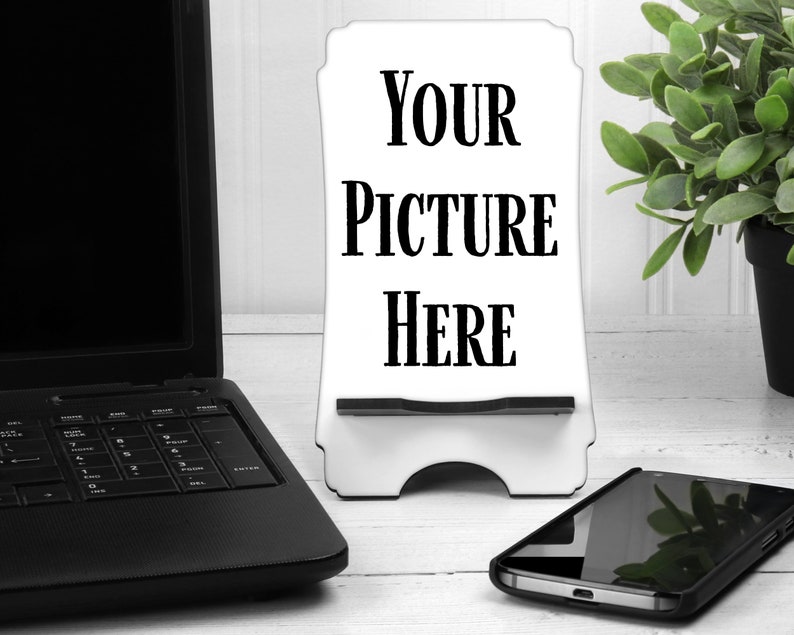 May include: White phone stand with the text "YOUR PICTURE HERE" on it. The stand is on a white wooden surface with a laptop and a smartphone next to it.