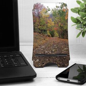 May include: A phone stand with a fall foliage design. The stand is made of wood and has a cutout for a phone. The background is a blurry image of a forest path.