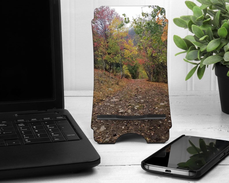 May include: A phone stand with a fall foliage design. The stand is made of wood and has a cutout for a phone. The background is a blurry image of a forest path.