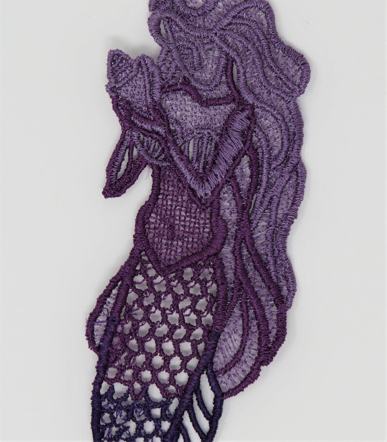 May include: Purple lace applique with a mermaid design. The lace is a gradient of purple shades, with a darker purple at the bottom and a lighter purple at the top.