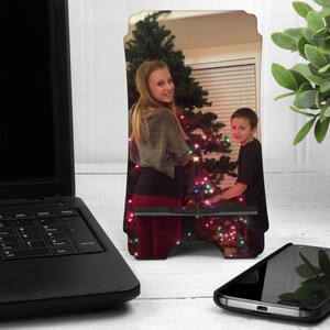 May include: A personalized phone stand with a photo of two people decorating a Christmas tree. The stand is made of wood and has a black base.