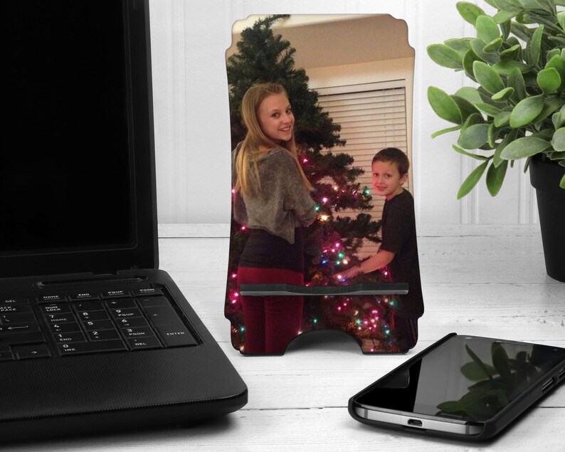 May include: A personalized phone stand with a photo of two people decorating a Christmas tree. The stand is made of wood and has a black base.