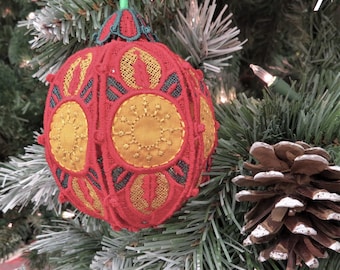 May include: Red Christmas ornament with gold and green embroidered details, hanging on a green Christmas tree. The ornament has a round shape with a detailed, openwork design. A pine cone is also visible.