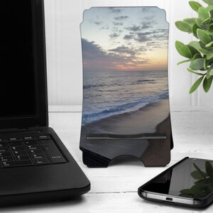 May include: A black phone stand with a sunset beach scene printed on it. The stand is made of metal and has a slot for holding a phone.