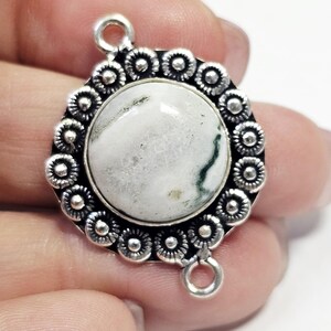 Puede incluir: Colgante plateado con una piedra central redonda y blanca con vetas verdes. La piedra está rodeada por un borde decorativo de pequeñas formas florales. El colgante tiene dos bucles para sujetarlo a una cadena u otra joya.
