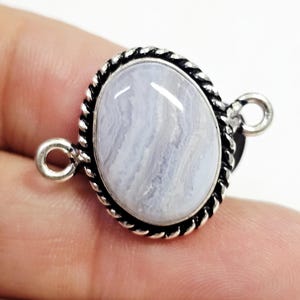 Puede incluir: Colgante plateado con una piedra preciosa ovalada de color azul claro. La piedra está engastada en un marco retorcido en forma de cuerda. El colgante tiene dos pequeños lazos para sujetarlo a una cadena. La piedra mide unos 2 cm de largo.