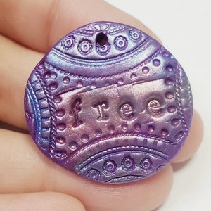 May include: A round, purple and iridescent clay charm with a hole at the top and the word "free" embossed on the front.