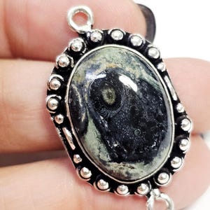 Puede incluir: Colgante ovalado con una piedra oscura estampada engastada en un marco de metal plateado. La piedra presenta tonos de negro, gris y verde. El marco tiene un borde de cuentas y bucles para la fijación.