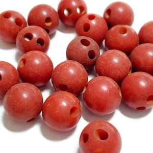 May include: A selection of red spherical beads, each with two or three holes. The beads are a deep reddish-brown hue and appear smooth. They are arranged on a white surface. The beads could be used for jewellery making or other craft projects.