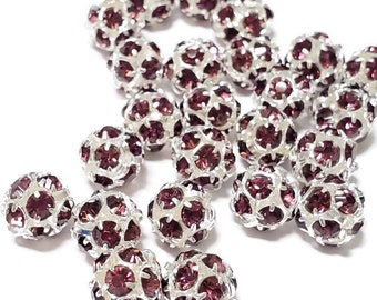May include: A scattering of round, silver-toned beads with a dark purple rhinestone design. The beads are scattered on a white surface.