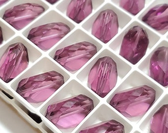 May include: A close-up shot of a white tray filled with faceted, elongated, purple-pink glass beads. Each bead is individually housed in a square compartment, showcasing their geometric cuts and color.