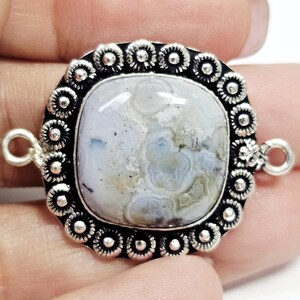 Puede incluir: Un colgante cuadrado plateado con un centro de piedra azul claro y gris. La piedra está enmarcada por un borde decorativo de pequeños acentos circulares. El colgante tiene dos bucles para sujetarlo a una cadena u otro componente de joyería.