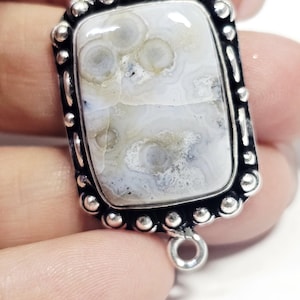Puede incluir: Un colgante rectangular con un marco plateado y una piedra de color claro. La piedra tiene un patrón único de remolinos blancos, grises y beige. El marco está adornado con pequeñas cuentas redondas plateadas. El colgante tiene bucles en la parte superior e inferior para una cadena.