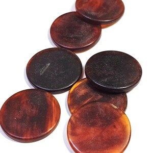 May include: A collection of seven circular buttons featuring a marbled, tortoiseshell-like pattern in varying shades of dark brown, amber, and black. Each button has a smooth, polished surface and a rounded edge, arranged in a loose cluster.