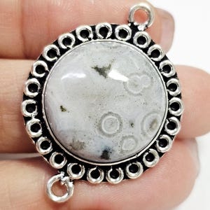 Puede incluir: Colgante redondo plateado con una piedra central gris claro y blanca. La piedra tiene un patrón único de círculos y motas. El colgante está rodeado por un borde decorativo y tiene dos bucles para sujetarlo.