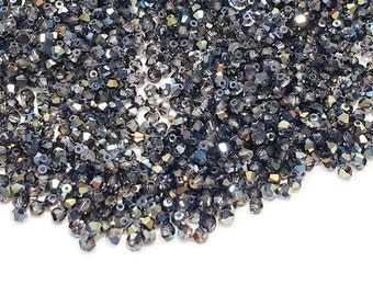 Puede incluir: Primer plano de un montón de pequeñas cuentas facetadas. Las cuentas son de un color gris oscuro iridiscente, con toques de azul y dorado. Están esparcidas sobre una superficie blanca, creando un efecto brillante. Ideal para la fabricación de joyas.