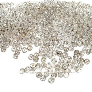 Op de afbeelding: Een stapel heldere, gefacetteerde kristallen strass-steentjes. De kleine, ronde strass-steentjes hebben meerdere facetten die het licht vangen, waardoor een sprankelend effect ontstaat. Ze zijn verspreid over een witte ondergrond.
