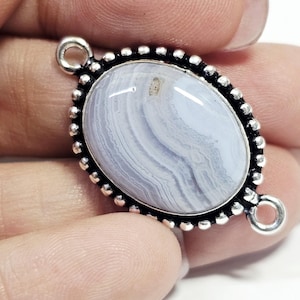 Puede incluir: Colgante ovalado de piedra preciosa de ágata de encaje azul con un borde plateado con cuentas y dos pequeños bucles. El ágata tiene un color azul claro con vetas blancas. El colgante está en una mano.