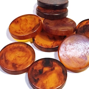 Puede incluir: Una colección de botones redondos y planos con un patrón jaspeado en tonos ámbar, marrón y naranja oscuro. Los botones tienen un acabado liso y brillante y están dispuestos en un montón disperso, mostrando sus variados patrones de colores.
