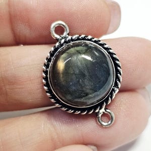 Puede incluir: Un colgante redondo plateado con una piedra oscura e iridiscente en el centro. La piedra está enmarcada por un borde retorcido en forma de cuerda. El colgante tiene dos pequeños bucles para sujetarlo a una cadena u otros componentes de joyería.