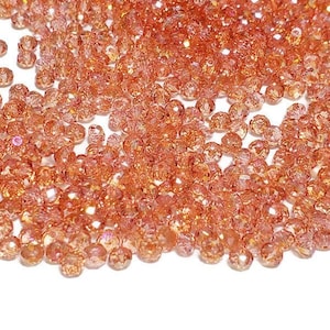 Op de afbeelding: Een grote hoeveelheid kleine, ronde, iriserende oranje glazen kralen. De kralen hebben een gefacetteerd oppervlak en zijn verspreid over een witte achtergrond.