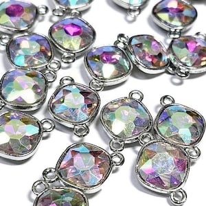 May include: A group of iridescent, faceted, square-shaped glass beads with silver-toned metal loops. The beads are arranged in a random pattern on a white surface.