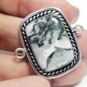 Puede incluir: Un colgante rectangular con un centro de piedra natural con patrones blancos y verdes. La piedra está engastada en un marco plateado con un borde trenzado y dos pequeños lazos para la fijación.
