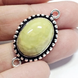 Puede incluir: Colgante ovalado con una piedra preciosa amarillo pálido, engastado en un marco de metal plateado. El marco tiene un borde con cuentas y dos pequeños bucles para sujetarlo a una cadena u otros componentes de joyería.