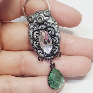 May include: A silver charm with a large clear crystal and a smaller green crystal hanging from it. The charm has a detailed design with black accents and a silver ring at the top.
