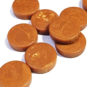 May include: A collection of circular, flat-topped buttons in a warm, medium-brown shade. Each button features a distinct, swirling marbled pattern in lighter tan and cream tones, creating a unique, organic appearance across the smooth surface.