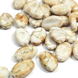 May include: A collection of oval-shaped beads in shades of cream and tan, with gold-colored vein-like accents. The beads have a glossy finish and are scattered on a white surface. These beads are suitable for jewelry making and crafting projects.