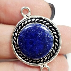 Puede incluir: Colgante redondo plateado con una piedra azul oscuro pulida en el centro. La piedra está rodeada por un borde decorativo trenzado. El colgante tiene dos pequeños lazos para sujetarlo a una cadena u otros componentes de joyería.