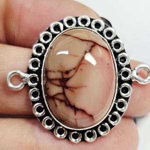 Puede incluir: Colgante ovalado con una piedra pulida de color marrón claro con vetas rojas oscuras. La piedra está engastada en un marco de metal plateado con un borde decorativo y dos bucles para la fijación. Primer plano.