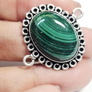 Puede incluir: Colgante ovalado de piedra malaquita con un borde decorativo plateado. La malaquita presenta patrones arremolinados de color verde oscuro y verde claro. El colgante tiene dos bucles para sujetarlo a una cadena u otro componente de joyería.