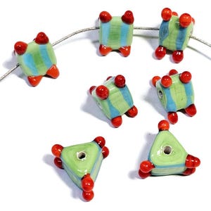 Puede incluir: Una colección de cuentas de cerámica hechas a mano en varias formas. Las cuentas presentan un patrón de rayas verdes y azules con detalles en rojo. Las cuentas están ensartadas en un alambre fino de color plateado, mostrando su diseño único y colores vibrantes.