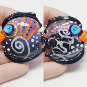 Puede incluir: Dos abalorios de pez de vidrio con detalles en negro, blanco, morado, naranja y azul. Los peces tienen un patrón de espiral y puntos blancos.