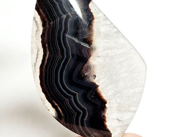 May include: A polished gemstone with a teardrop shape. The stone features layers of black and white bands, with a translucent white area on one side. The gemstone is held between two fingers.