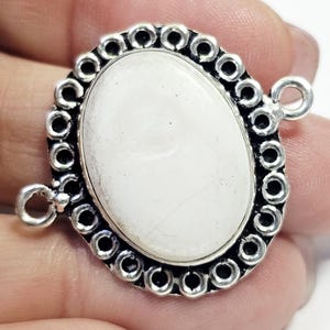 Puede incluir: Colgante ovalado de piedra blanca con un marco plateado adornado. El marco presenta una serie de pequeños diseños circulares alrededor del borde de la piedra. Dos bucles están unidos para colgar o conectar a otras joyas. Mide unos 3 cm.