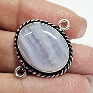 Puede incluir: Colgante ovalado con una piedra azul claro, engastada en un marco de metal plateado. El marco tiene un diseño de cuerda trenzada alrededor de la piedra e incluye dos pequeños bucles.