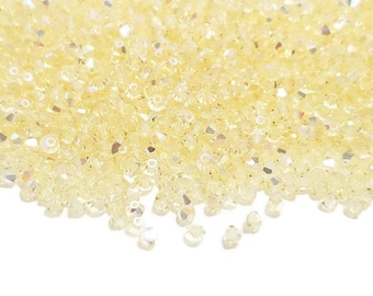Puede incluir: Primer plano de un montón de pequeñas cuentas de color amarillo claro con facetas iridiscentes. Las cuentas están dispersas, captando la luz y creando un efecto brillante. Las cuentas se utilizan probablemente para la fabricación de joyas u otros proyectos de artesanía.