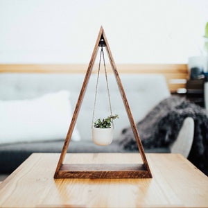 May include: A wooden triangular plant hanger with a small potted plant hanging from it. The hanger is sitting on a wooden table.