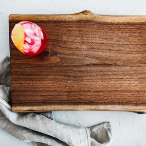 May include: A red cocktail with a lemon wedge garnish sits on a wooden chopping board. The board has a natural edge and is made of dark wood.