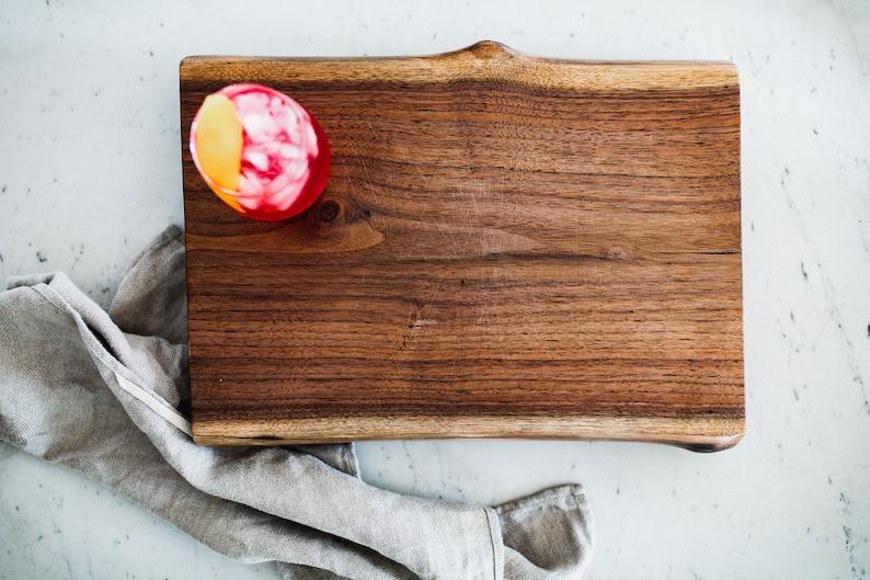 May include: A red cocktail with a lemon wedge garnish sits on a wooden chopping board. The board has a natural edge and is made of dark wood.