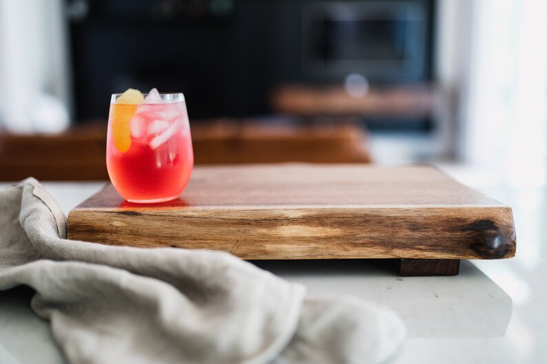 May include: A glass of red cocktail with ice and a lemon wedge on a wooden serving board.