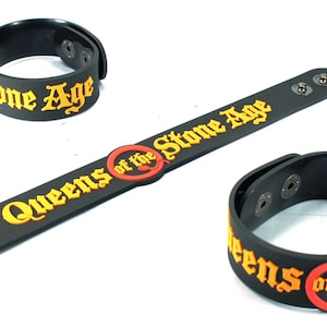 May include: Black wristbands with the text "Stone Age" and "Queens of the Stone Age" in yellow lettering. One long band and two shorter bands are shown. The band has a snap closure.