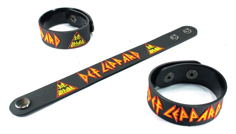 May include: Black Def Leppard wristbands with the band's logo in orange and yellow. The wristbands are made of a flexible material and have snap closures. The image shows three wristbands, one long and two circular.