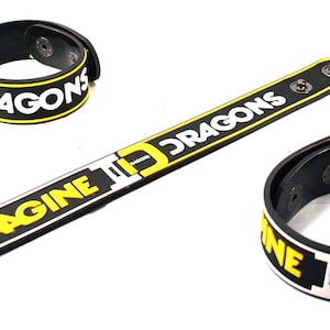 Puede incluir: Tres pulseras de silicona negras y amarillas con el texto "Imagine Dragons" y "Dragons" impreso en ellas.