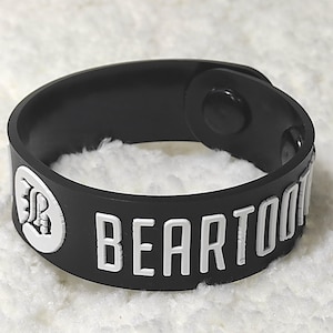 Puede incluir: Pulsera de goma negra con la palabra "BEARTOOTH" en letras blancas. La pulsera tiene un cierre de botón a presión y un logotipo blanco. La pulsera mide aproximadamente 2,5 cm de ancho.