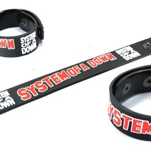 Könnte beinhalten: Schwarze Armbänder und ein langes Armband mit dem Text "SYSTEM OF A DOWN" in Rot und Weiß. Die Armbänder haben Druckknöpfe. Das Armband hat Druckknöpfe und einen einzelnen Druckknopf.