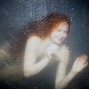 May include: A woman with long red hair is partially submerged in water. Her face is visible, and she is looking down. The water is clear and the background is a dark blue.